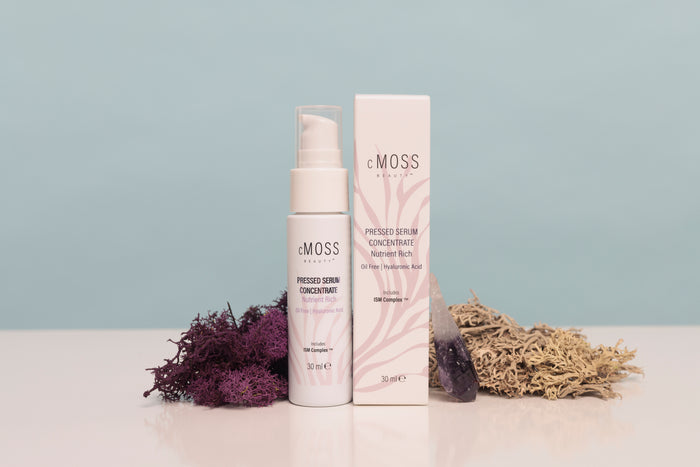 eMOSS Protected Serum Concentrate shown with its white pump bottle and matching rectangular box on a white surface, styled with dried purple and beige botanicals against a pale blue background.