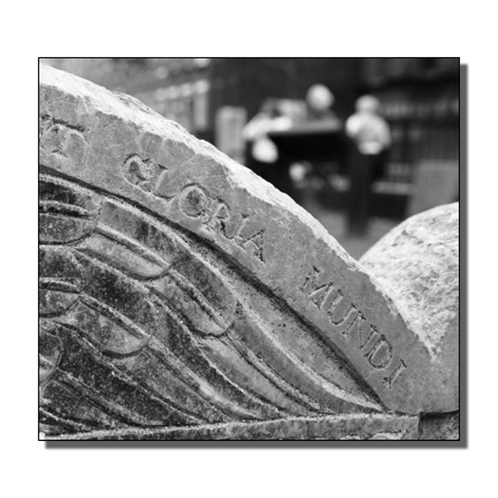 A close-up of an old stone monument engraved with Gloria Mundi; in the blurred background, two people stand by a building. This black and white image would make a striking cover for a Ghost Thrower music album or 10-song CD.