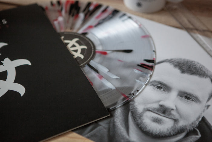 A clear vinyl record with black and red streaks, partly in a black sleeve with a white geometric logo, sits next to a grayscale photo of Brian Marquis. This limited Blood And Spirits pressing by Brian Marquis rests on a wooden surface.