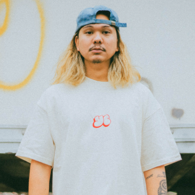 A person with long blond hair, wearing a stylish backwards blue cap and a Young Culture T-shirt with a red cloud design, stands outside in front of a graffiti-covered wall.