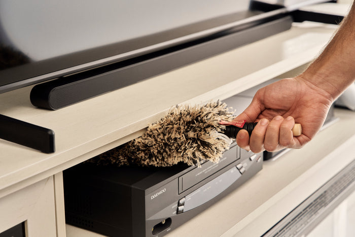 A hand holding a fluffy duster cleans a black Quincy DVD player beneath a TV on a cream-colored media console.
