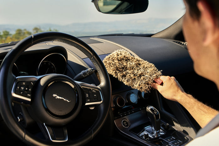 A person inside a car uses a fluffy beige microfiber duster on the dashboard, with a black Jaguar steering wheel and center console visible in the background.