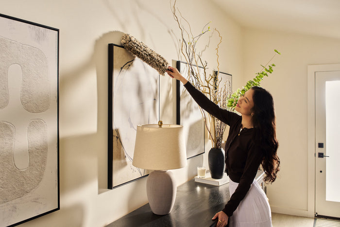 A woman in a dark brown long-sleeve top and white skirt reaches up to adjust a tall dried floral arrangement hanging on a cream-colored wall in a modern home interior.