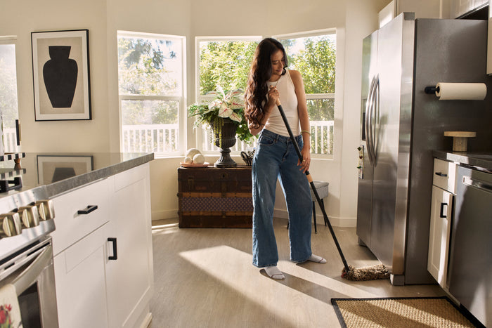 A woman mopping a light wood floor in a bright, modern kitchen with stainless steel appliances and white cabinets.