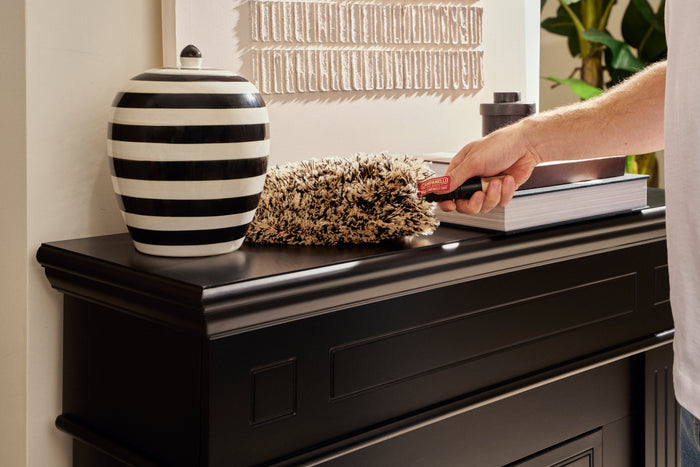 A close-up lifestyle shot of a person's hand using a black-handled duster to clean a dark wood console table. The table is styled with a black and white striped ceramic jar and a textured beige throw blanket, set against a warm beige wall with a woven wall hanging.