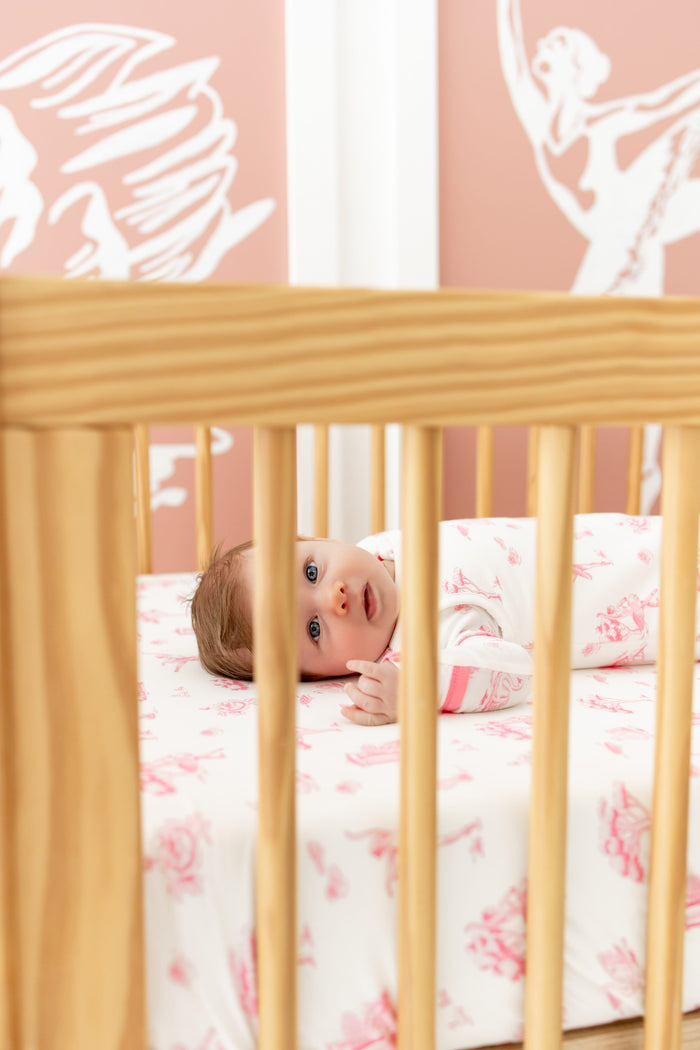 Baby laying on a crib on top of the Crib Sheet in Toile Ballet wearing the sleep bag and matching romper