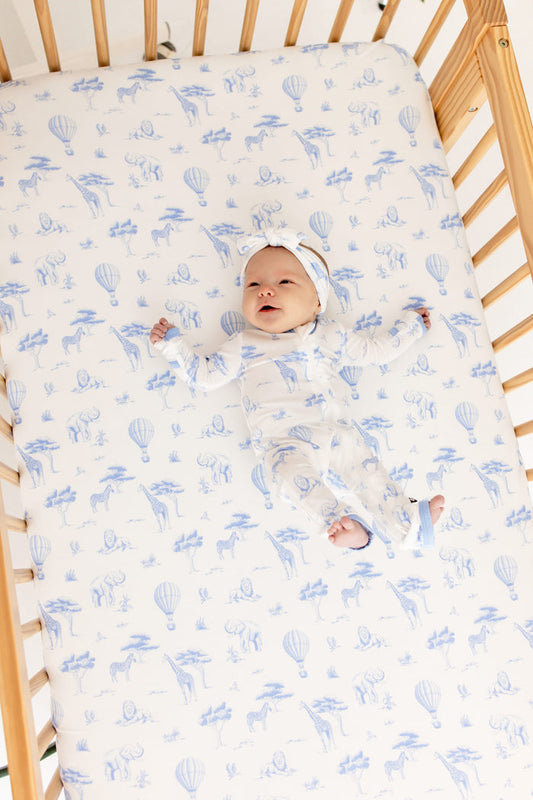 Infant laying in a crib on a Crib Sheet in Toile Safari wearing the matching zippered romper and bow