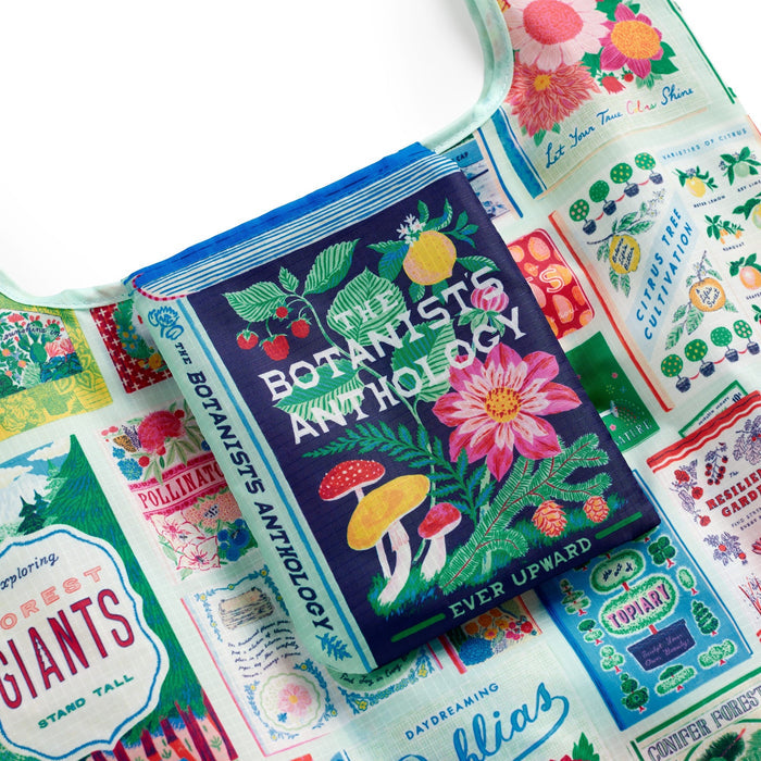 The Emily Taylor Ever Upward Botanist Bookshelf Packable Nylon Tote Bag, featuring vibrant botanical and inspirational designs, is shown with a colorful book illustrated with flowers, mushrooms, and plants.