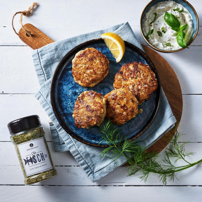 Fischfrikadellen mit Kräuterdip und Zitrone auf Teller, daneben Fischgewürz und frischer Dill auf hellem Tisch.
