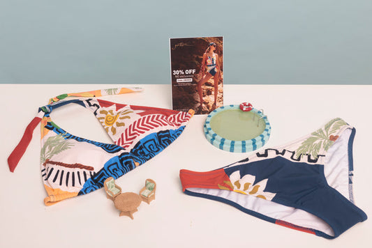 A coordinated swimwear set laid out on a white surface against a pale blue wall, including a multicolor bikini top with red, blue, yellow, and white tropical/geometric patterns, matching bikini bottoms in navy, white, and red with a floral print, a small round green float, and a small wooden float.