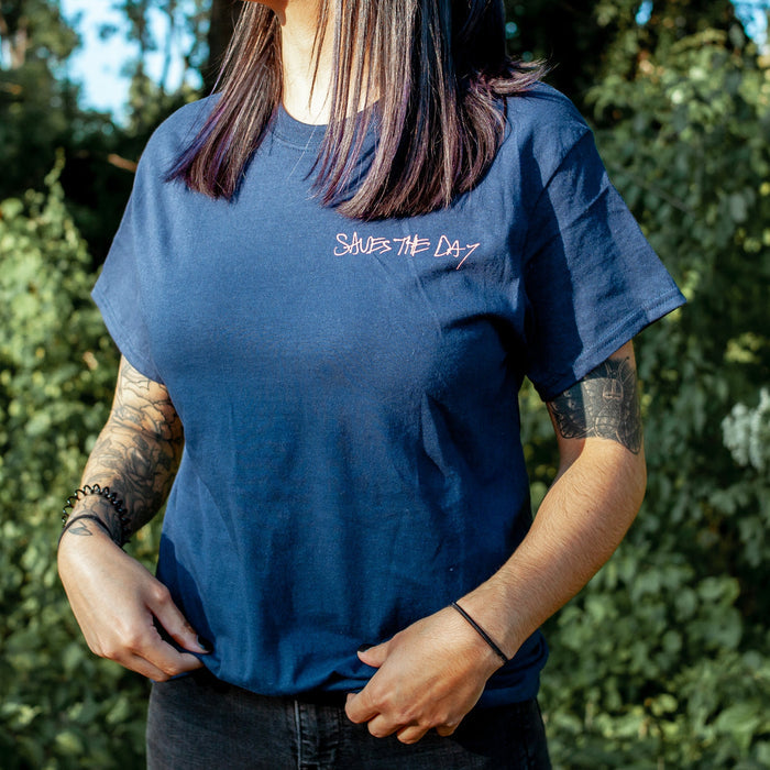 A person with dark hair and tattooed arms stands outdoors wearing a Saves The Day blue T-shirt with “SAVES THE DAY” in white script, surrounded by greenery and sunlight—a must-have design for fans.
