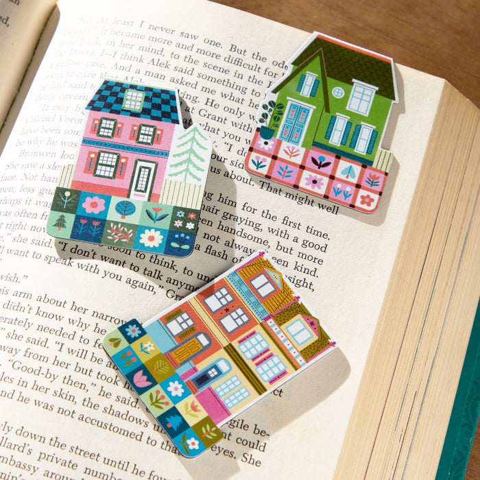 Three Home Sweet Home Magnetic Bookmarks by Molly Egan, each with a colorful house-shaped floral design, rest on the open pages of a book in bright sunlight.