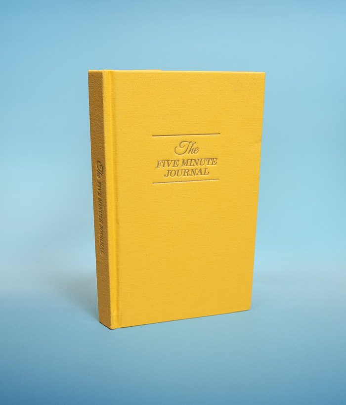 Yellow journal titled 'The Five Minute Journal' on a light blue background