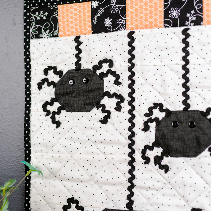 A close-up of a Halloween-themed quilted textile featuring two black spider appliqués on a white dotted background, with black-and-white polka dot trim, orange and black patterned fabric accents, and a dark gray wall behind it.