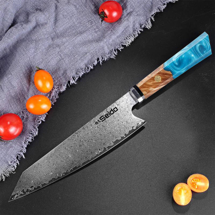 A Sado Japanese kitchen knife with a Damascus steel blade and a colorful wood-and-blue handle, styled diagonally on a dark surface with cherry tomatoes and a gray cloth.