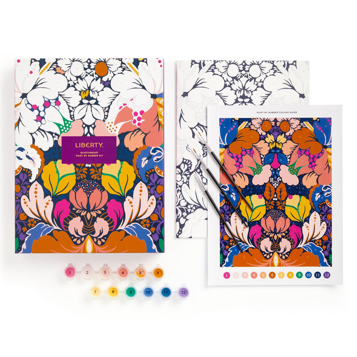 The Liberty London 9 x 12 Paint by Number Kit Glastonbury features a floral design, numbered premium acrylic paints, and two brushes in a box showing a partly finished painting, all arranged on a white background.