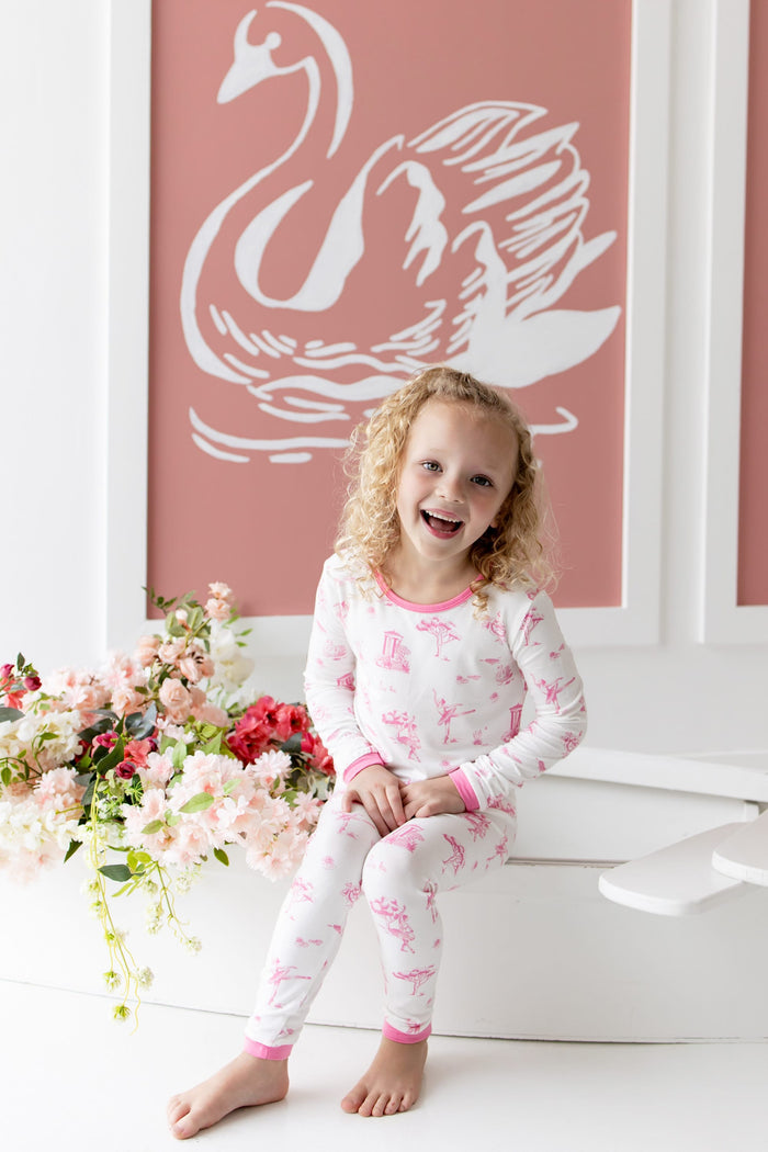 Young smiling girl wearing the Long Sleeve Pajamas in Toile Ballet sitting on the edge of a white row boat beside flowers in front of a white swan on pink background