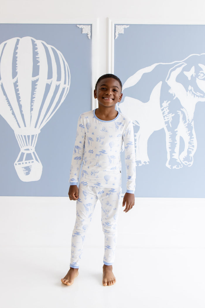 Smiling boy wearing the Long Sleeve Pajamas in Toile Safari standing in front of safari inspired blue and white paintings