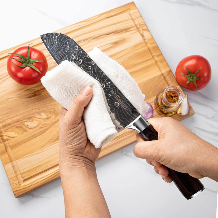Person cleaning a chef's knife with a white cloth on a wooden cutting board with tomatoes and spices.
