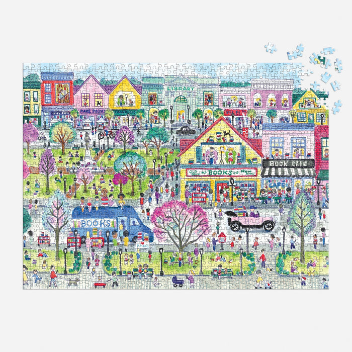 The Michael Storrings Book Town 1000 Piece Puzzle by Michael Storrings features a vibrant town with people, a bookshop, book café, book bus, pink-blossomed trees, vintage cars, pastel buildings, and unfinished pieces in the top right corner.