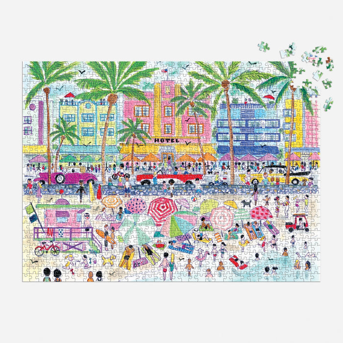 The Michael Storrings South Beach Miami 1000 Piece Puzzle by Michael Storrings features a vibrant beach scene with people, umbrellas, palm trees, and art deco buildings. A few pieces are missing in the top right corner.