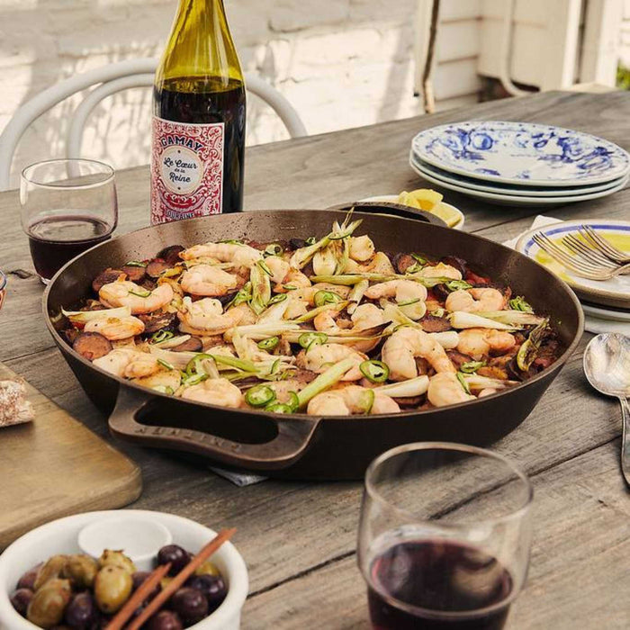 Smithey No. 14 dual handle skillet on an outdoor dining table filled with plates, wine glasses and a bottle of wine for al fresco dining