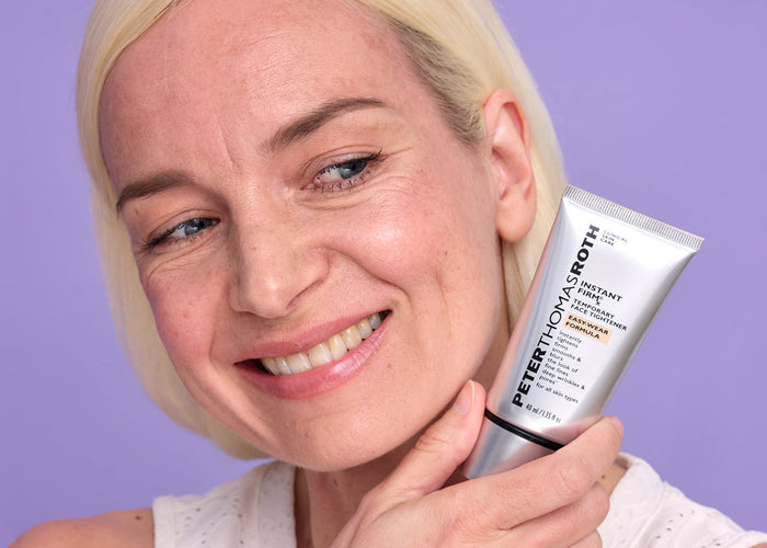 A smiling blonde woman holds a white cosmetic tube against a solid lavender-purple background. The tube is labeled Estérieux Smooth.