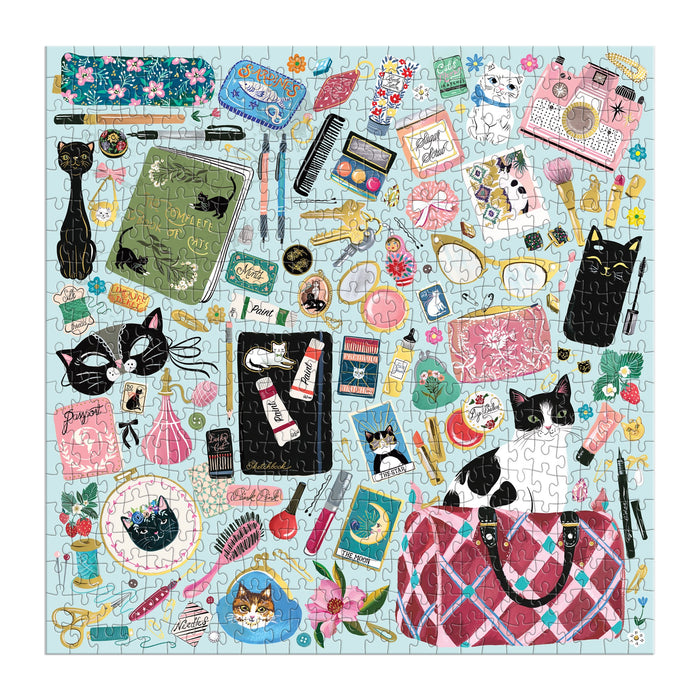 The Christine De Carvalho Purrfect Accessories 500 Piece Foil Puzzle features a vibrant collage of cat-themed items—like stationery, makeup, figurines, plants, and cute feline illustrations—on a light background.