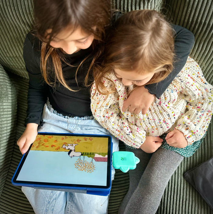 Two children sitting closely together on a green corduroy couch, looking at a tablet screen. One child holds a blue tablet case, and the other wears a cream knit sweater with colorful flecks.