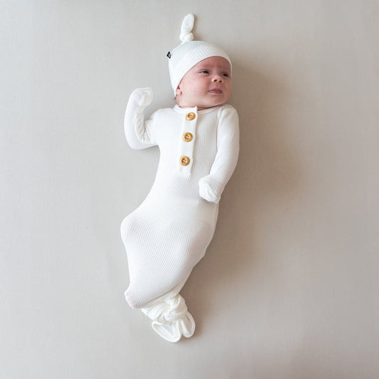 Baby laying on eggshell surface wearing Kyte Baby Ribbed Knotted Gown with Hat Set in Cloud. Baby is looking to the upper right-hand corner