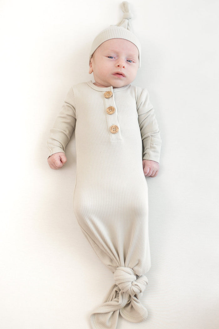 Baby in laying wearing the Kyte Baby Ribbed Knotted Gown with matching hat in the color oat.