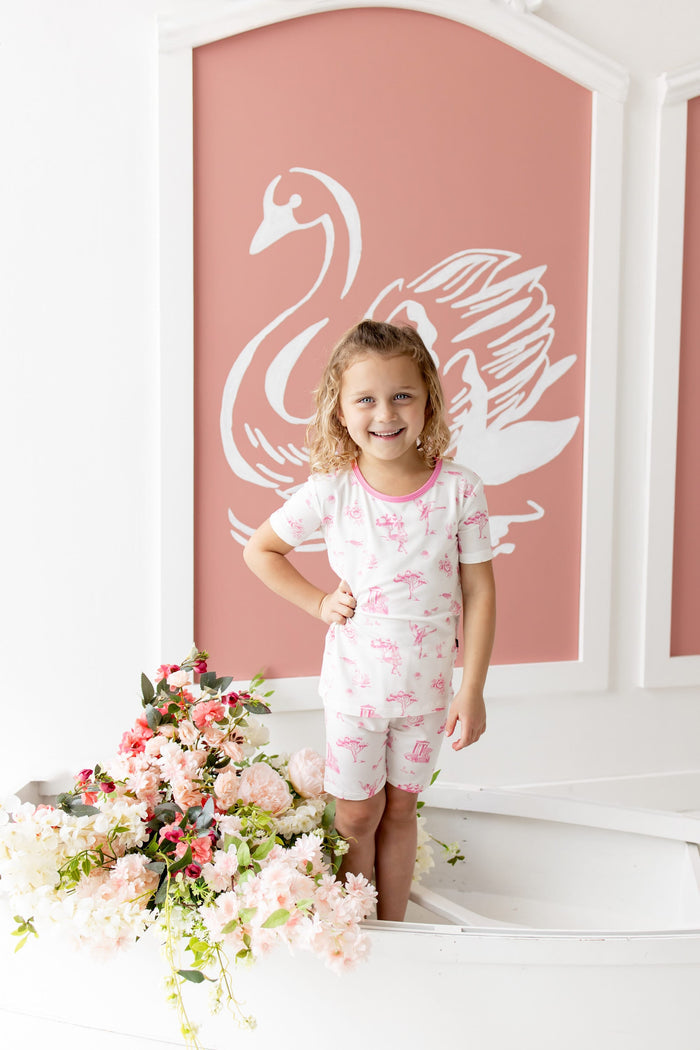Young girl wearing the Short Sleeve Pajamas in Toile Ballet posing standing inside a white rowboat beside flowers in front of a white swan on a pink background painting