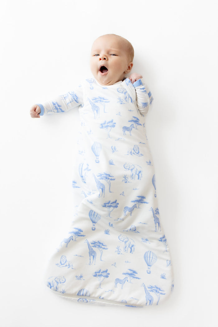 Yawning infant laying on a white surface wearing the Sleep Bag in Toile Safari 1.0 and matching romper