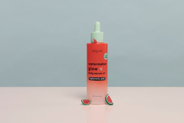 A bottle of SPALiPS Watermelon Glow Daily Serum Oil with a pink-to-white gradient body, a mint green dropper cap, and watermelon slice props on a light pink surface against a gray background.