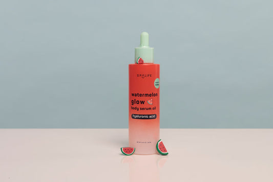 A bottle of SPALiPS Watermelon Glow Daily Serum Oil with a pink-to-white gradient body, a mint green dropper cap, and watermelon slice props on a light pink surface against a gray background.