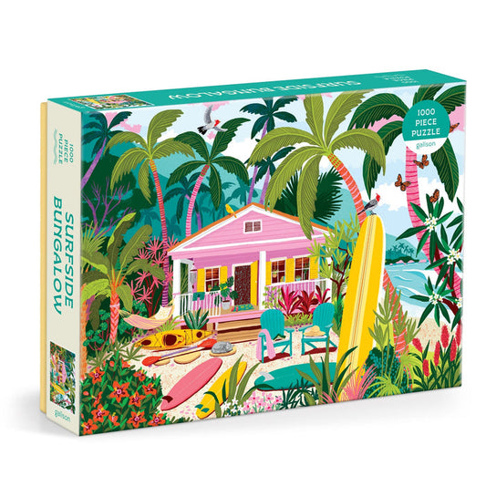 The Julia Cornelius Surfside Bungalow 1000 Piece Puzzle features a tropical beach scene with a pink bungalow, palm trees, surfboards, kayaks, and beach chairs by the ocean under a bright blue sky.