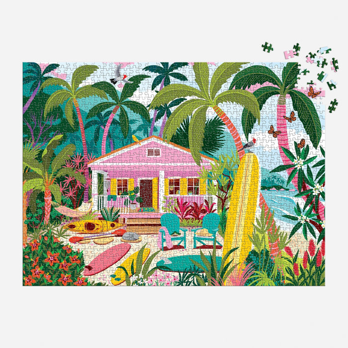 The Surfside Bungalow 1000 Piece Puzzle by Julia Cornelius features a vibrant tropical beach with a pink house, palm trees, surfboards, chairs, and lush plants. The top right corner shows some unfinished pieces for an added challenge.