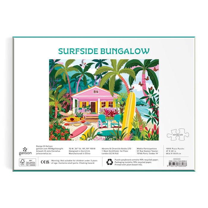 The image shows the back of a Julia Cornelius Surfside Bungalow 1000 Piece Puzzle box featuring a vibrant illustration of a pink house with palm trees, tropical plants, a pool, and surfboard. Product details are printed below the artwork.
