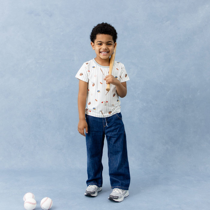 Young boy wearing the Toddler Basic Tee in Home Run and dark wash jeans holding a miniature baseball standing behind baseballs