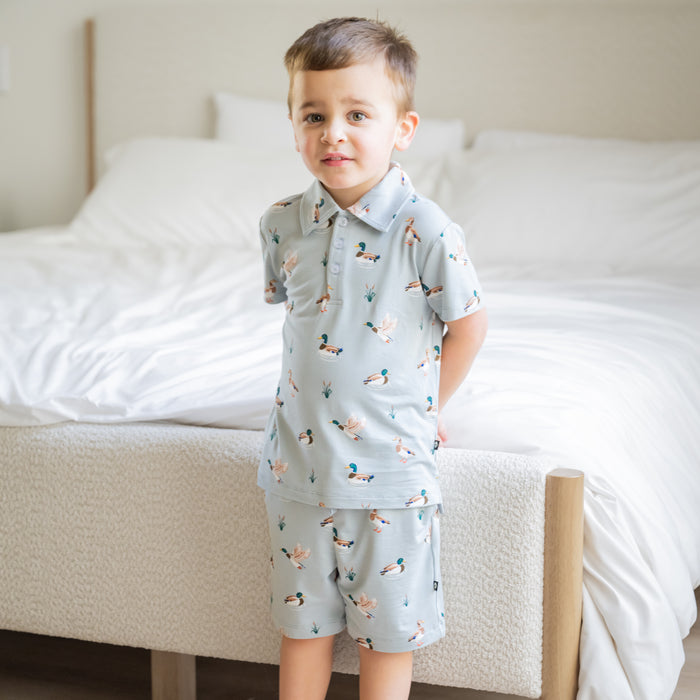 Young boy standing at the end of a bed wearing the Toddler Short Sleeve Polo in Mallard Duck with matching drawstring shorts