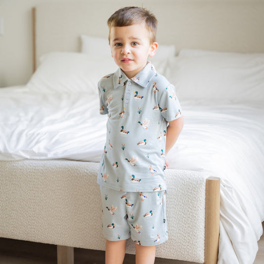 Young boy standing at the end of a bed wearing the Toddler Short Sleeve Polo in Mallard Duck with matching drawstring shorts