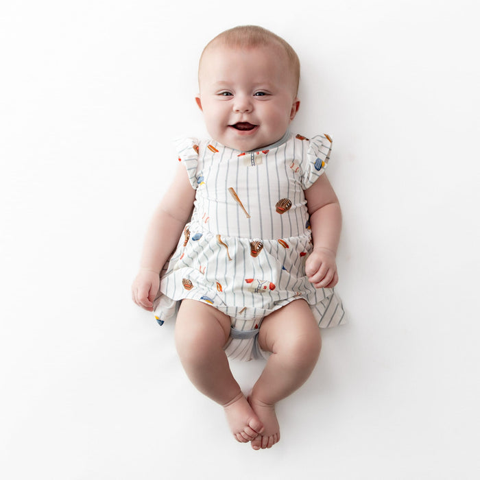 Smiling infant wearing the Twirl Bodysuit Dress in Home Run laying on a white surface