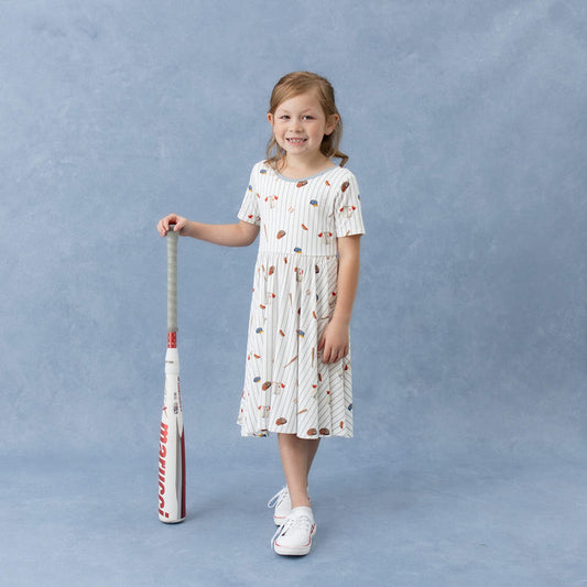Young girl wearing the Twirl Dress in Home Run standing beside a baseball bat