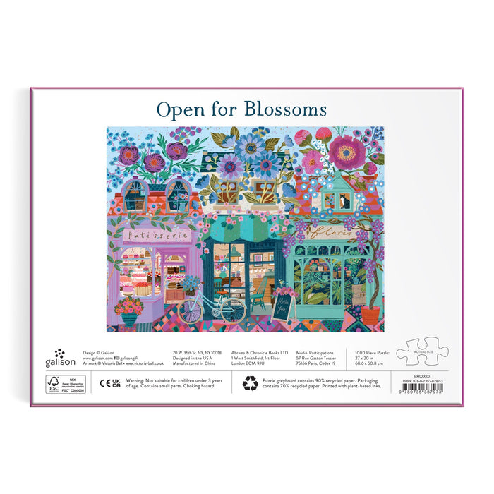 The Victoria Ball Open For Blossoms 1000 Piece Puzzle features a colorful illustration of three charming floral shops with vibrant flowers on their rooftops and storefronts, and “Open for Blossoms” on the box.