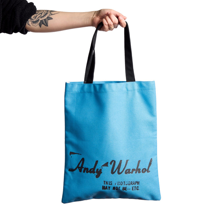 A person with a tattooed forearm holds up the Warhol Soup Can Volume 3 Canvas Tote Bag by Andy Warhol Foundation For The Visual Arts—a light blue tote with black handles and text on a white background.