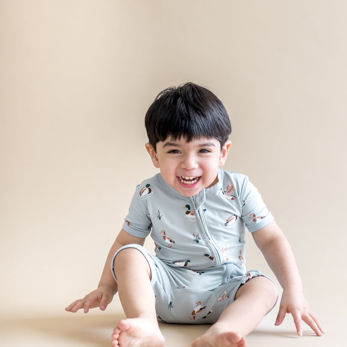 Young laughing boy sitting on the floor wearing the Zipper Shortall Romper in Mallard Duck
