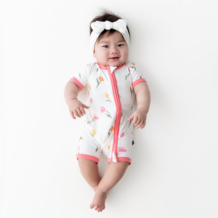 Young infant wearing the Zipper Shortall Romper in Tulip paired with a white bow headband