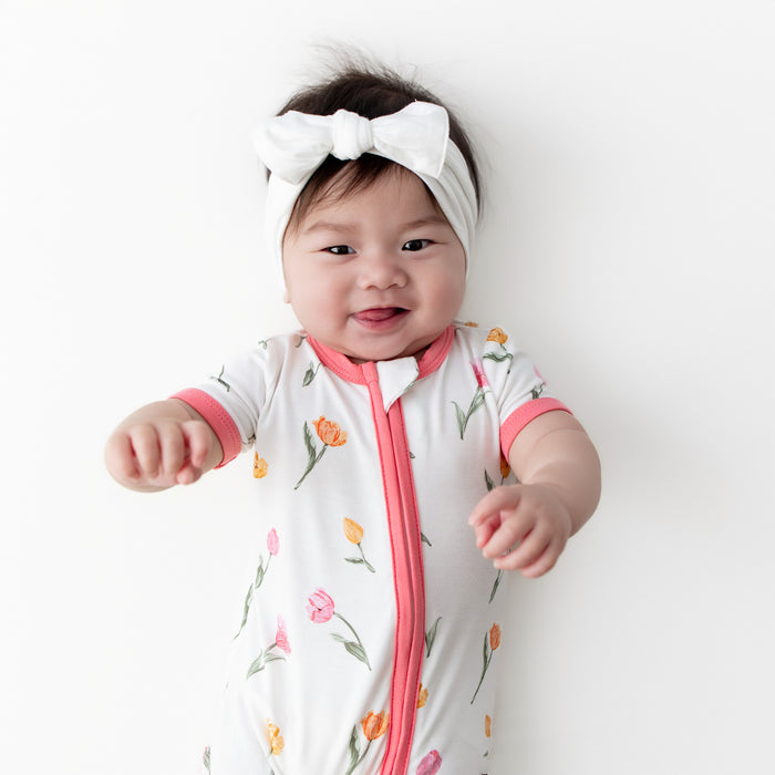 Close up of the Zipper Shortall Romper in Tulip shown on an infant model wearing a white bow