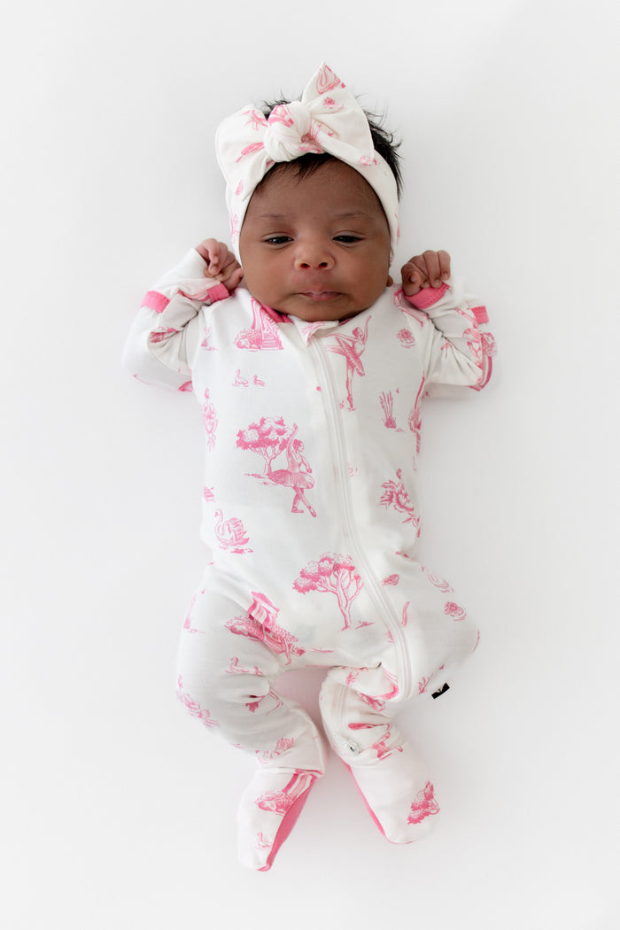 Newborn baby in a white and pink toile print footed pajama with a matching headband on a white background