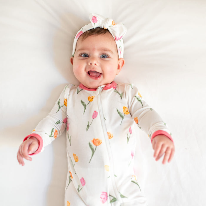 Close up of a toddler laying on a bed wearing the Zippered Romper in Tulip with matching knotted bow headband
